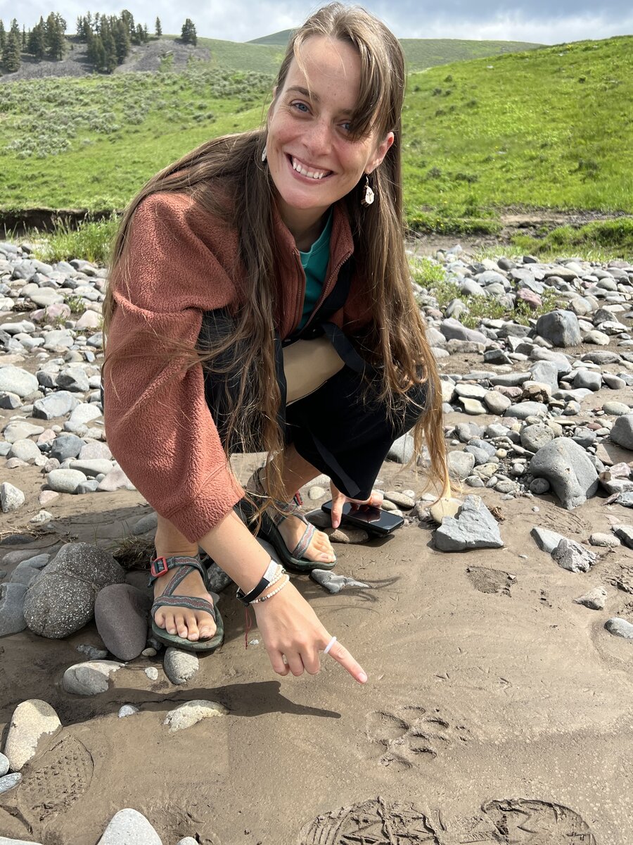 Emily kneeling by a riverbank pointing at animal tracks in the mud